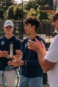 Junior Ryker Fringer, Senior AJ Wilkinson, and Coach Jason Enevoldson clap on the tennis court. 